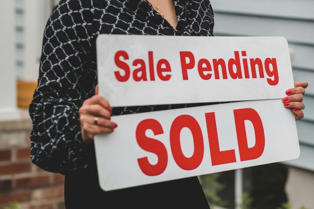 Person holding a real estate sign reading “Sale Pending” above “Sold” in front of a residential home.