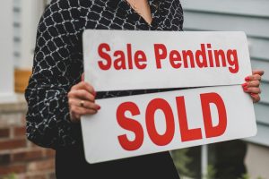 Person holding a real estate sign reading “Sale Pending” above “Sold” in front of a residential home.